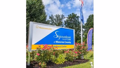 Outdoor view of a sign for Signature HealthCARE of Monroe County Rehab & Wellness Center surrounded by flowers and greenery, with a flagpole and blue sky in the background.