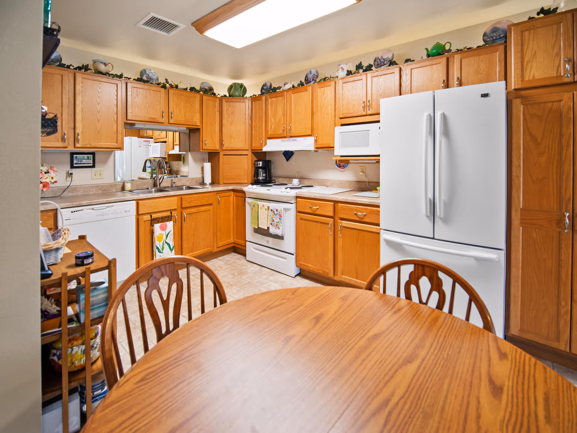 A kitchen with wooden cabinets, a white refrigerator, stove, microwave, and dishwasher. There is a wooden dining table with chairs in the foreground. Decorative plates and greenery are displayed above the cabinets.