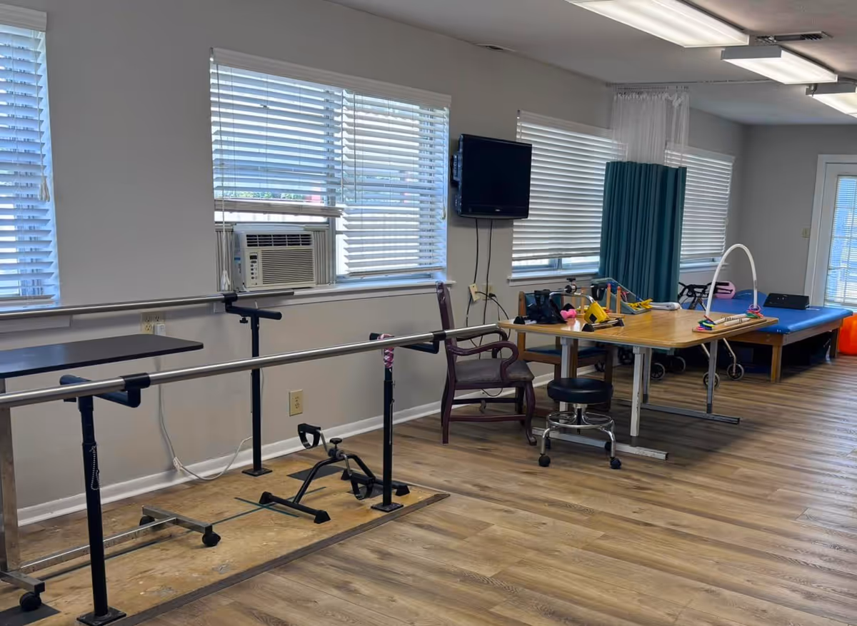 A rehabilitation room with parallel bars for walking exercises, a pedal exerciser on the floor, a table with various therapy tools, a chair, a stool, a therapy bed, and a walker. The room has large windows with blinds, a wall-mounted TV, and wood-patterned flooring.