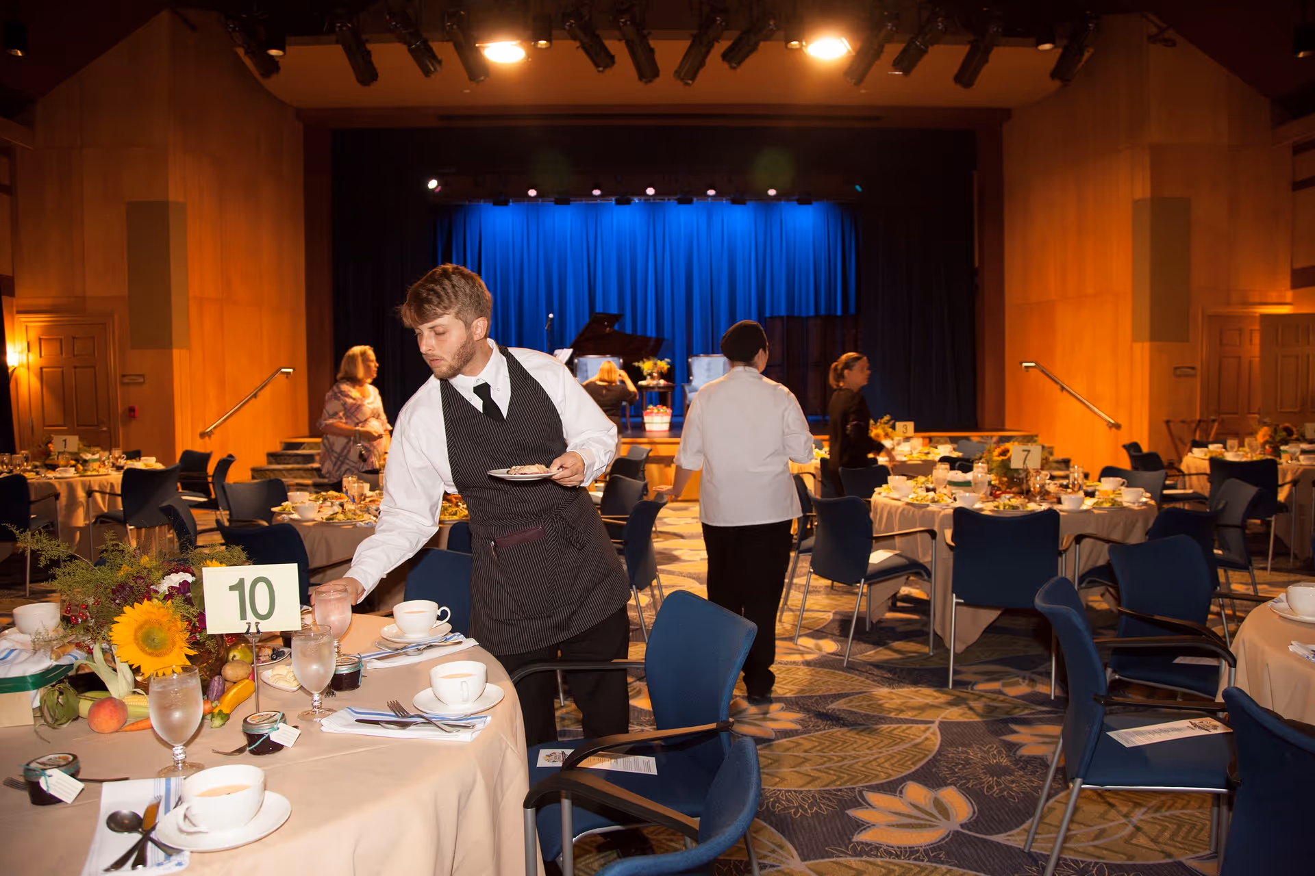 A banquet hall with round tables set for a meal, decorated with flowers and table numbers. Several waitstaff are attending to the tables, and a stage with a blue curtain and a piano is visible in the background.
