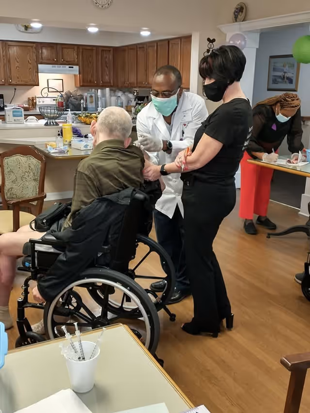 A healthcare professional wearing a white coat and face mask is administering a vaccine or injection to an elderly man seated in a wheelchair. A woman wearing black clothes and a face mask stands nearby, holding the elderly man's arm for support. In the background, another woman wearing a face mask is writing on a clipboard at a table in a room with wooden flooring and kitchen cabinets.
