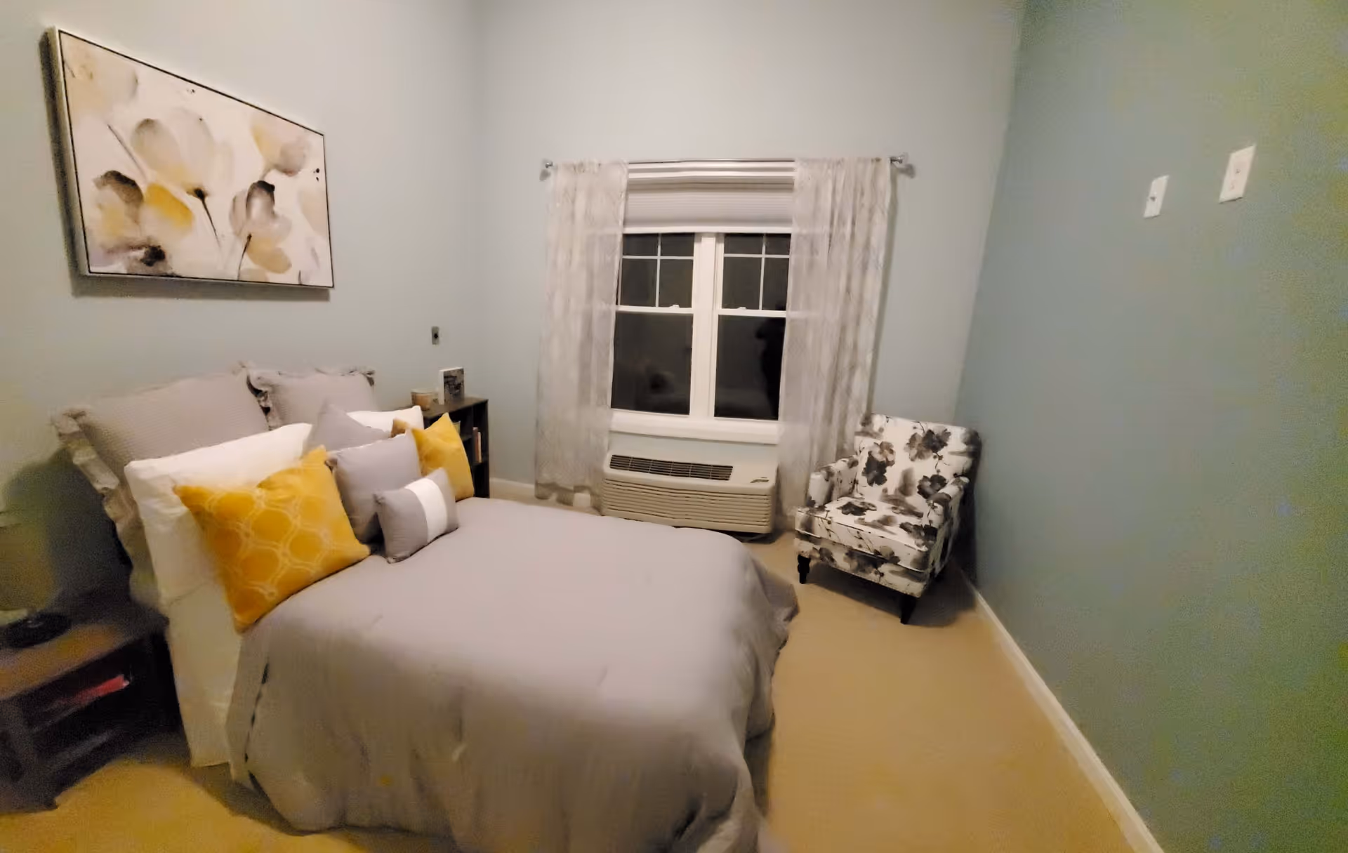 A cozy bedroom with a neatly made bed featuring gray bedding and multiple pillows in white, gray, and yellow. There is a floral patterned armchair in the corner, a window with sheer curtains above an air conditioning unit, and a piece of abstract floral artwork on the wall above the bed.