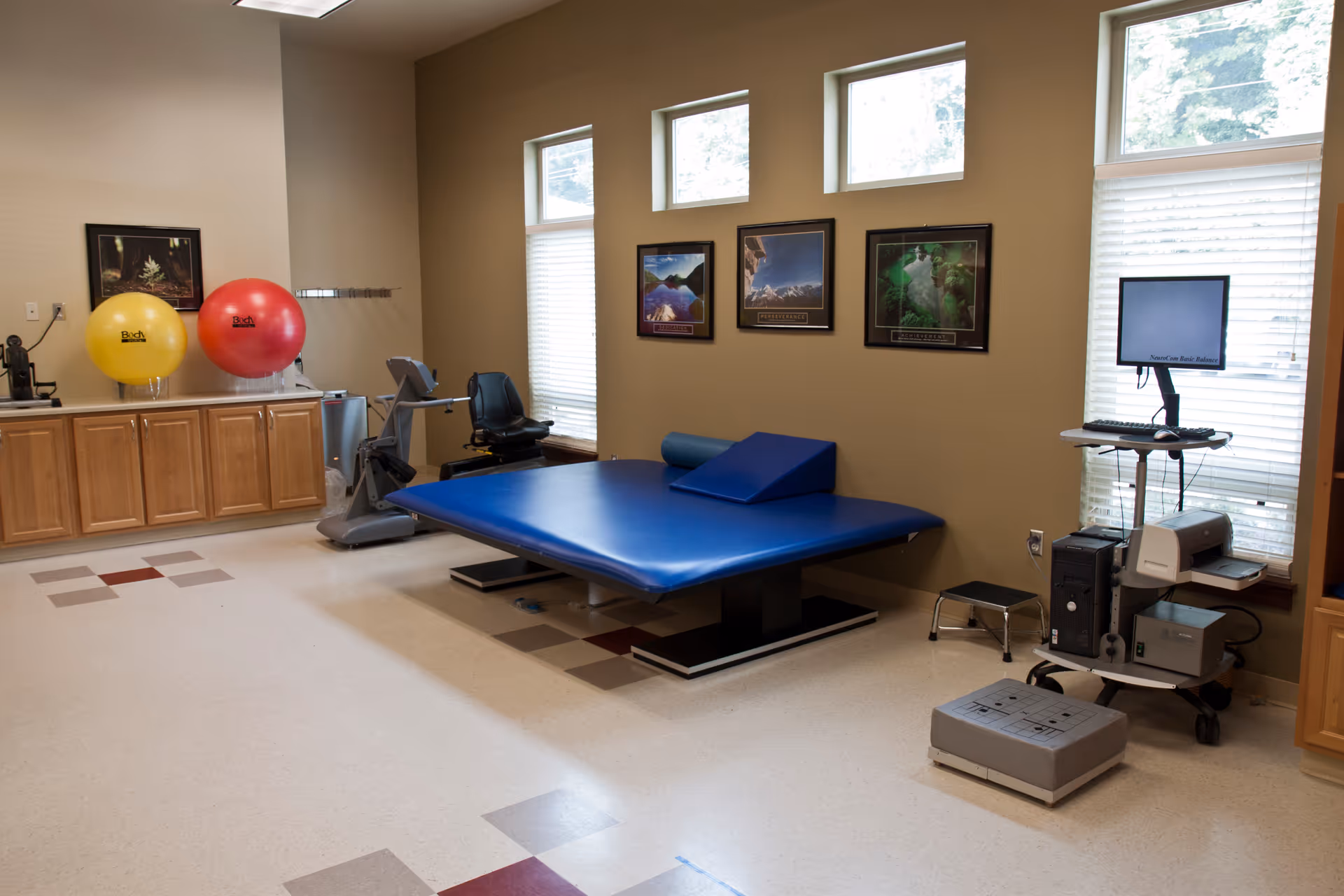 A rehabilitation or physical therapy room with a large blue padded therapy table, exercise equipment including a stationary bike, two large exercise balls on a wooden cabinet, a computer workstation with a monitor and printer, and three framed motivational posters on the wall. The room has beige walls, windows with blinds, and a tiled floor with a pattern of red and gray squares.