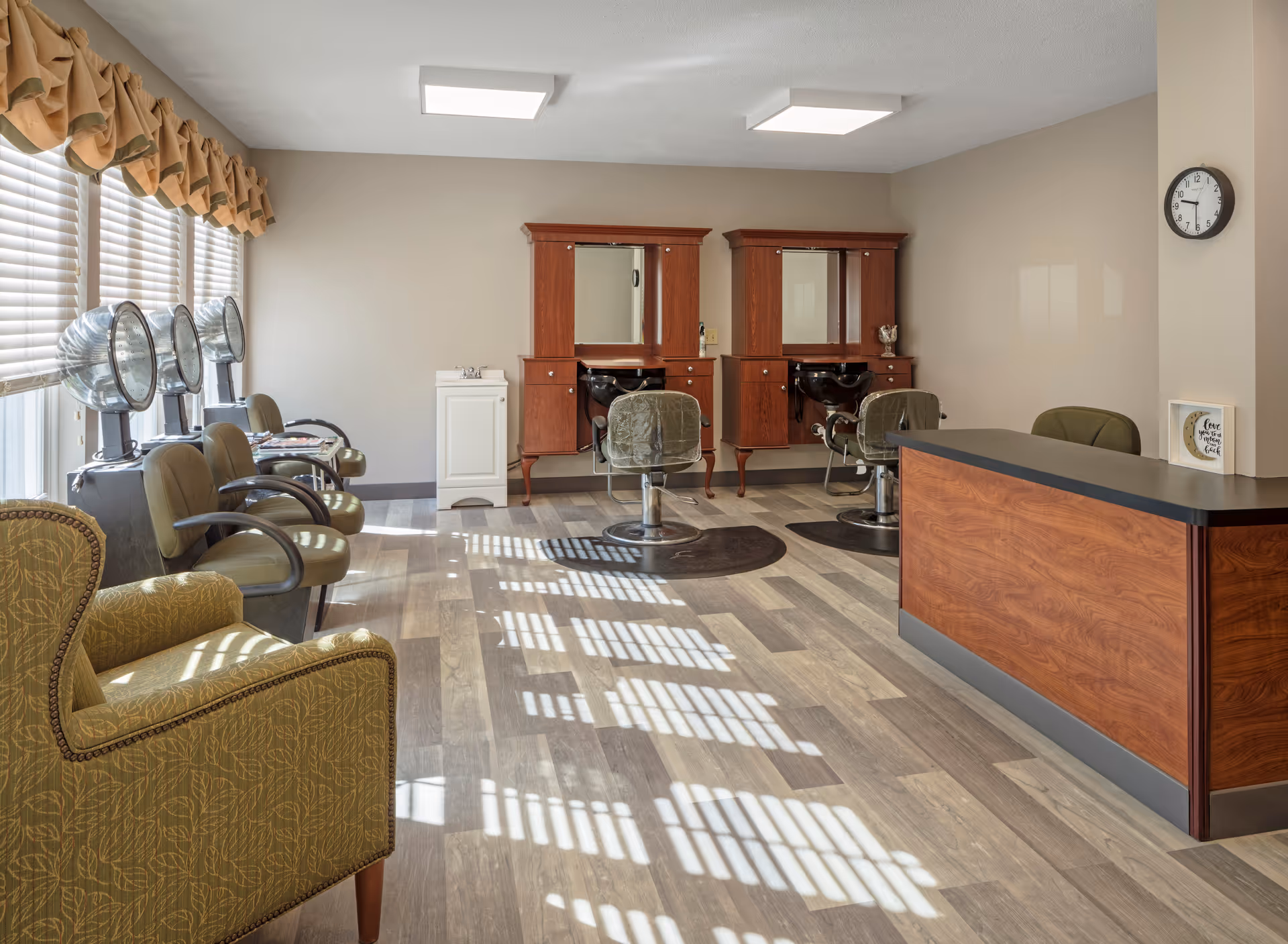 Interior view of a hair salon area in a senior living facility with two styling stations featuring mirrors and sinks, several salon chairs, hair dryers by the window, a reception desk, and a clock on the wall.