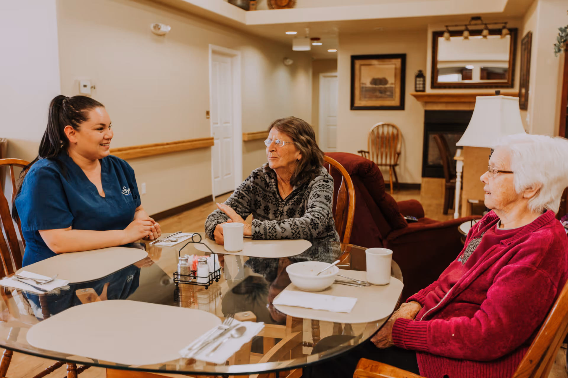 A caregiver in blue scrubs sits at a glass dining table with two elderly women, engaging in conversation. The table has placemats, utensils, cups, and a bowl. The room has warm lighting, wooden chairs, and a cozy interior with framed artwork and a fireplace in the background.