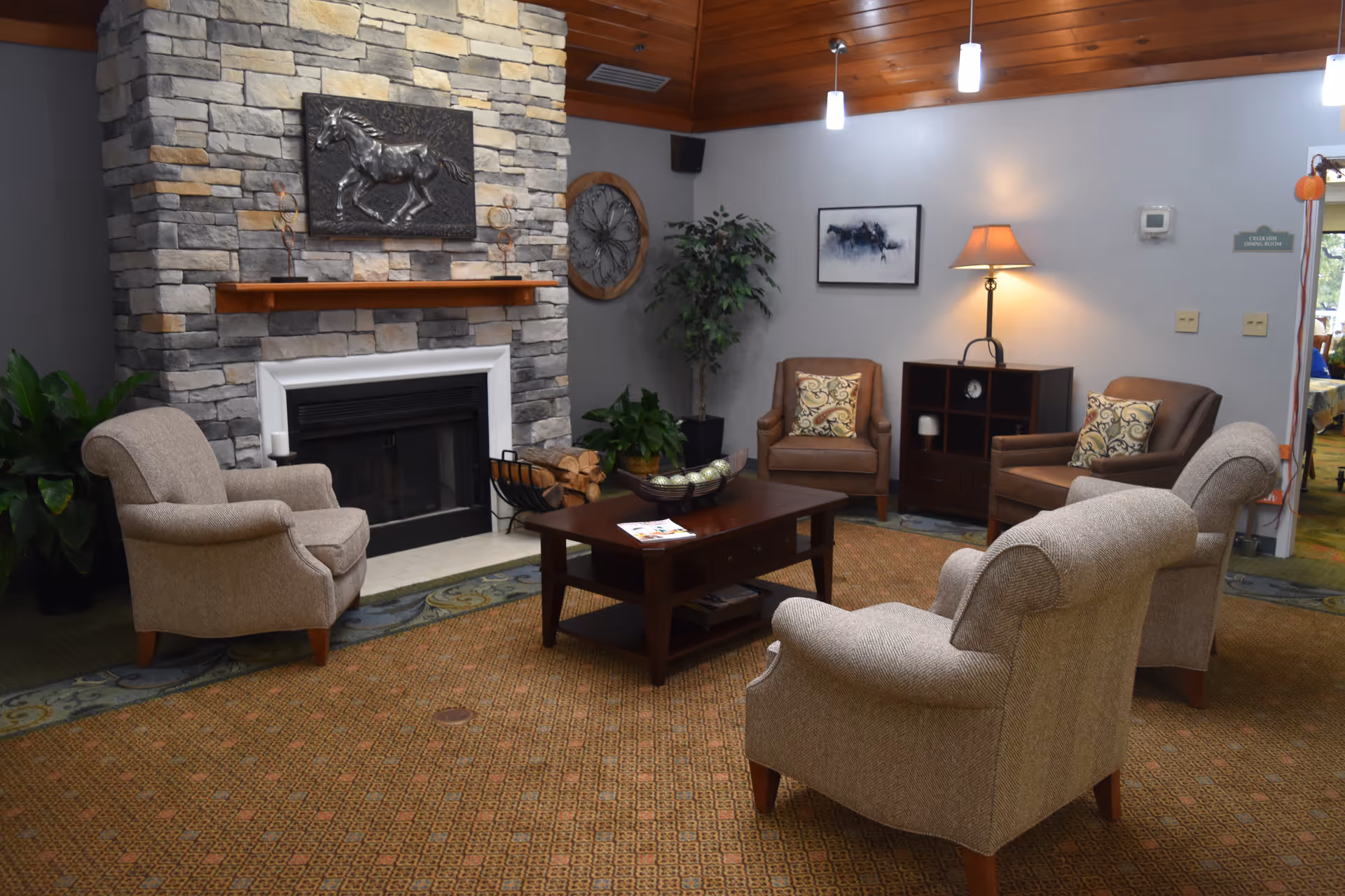 A cozy senior living common area with a stone fireplace featuring a horse sculpture above the mantel. The room has four upholstered armchairs arranged around a wooden coffee table with decorative items and magazines. There is a floor lamp, a small cabinet, wall art, and potted plants. The ceiling is wooden with hanging pendant lights.