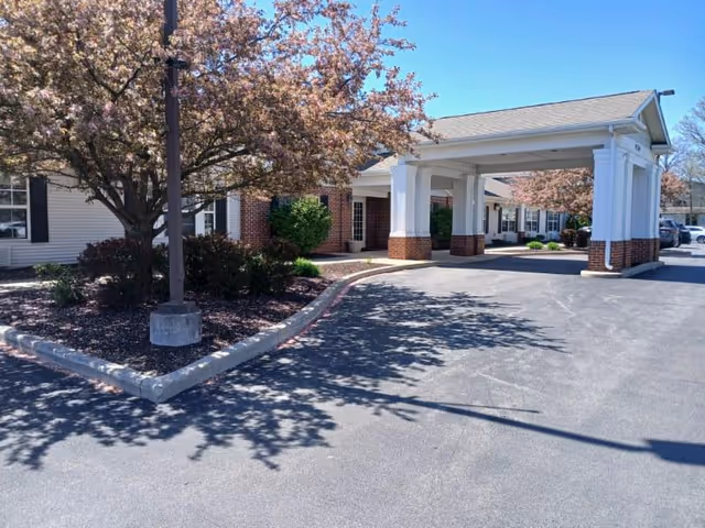 Exterior view of a senior living facility entrance with a covered drop-off area supported by white columns with brick bases. There are blooming trees and shrubs along the driveway, and a clear blue sky overhead.