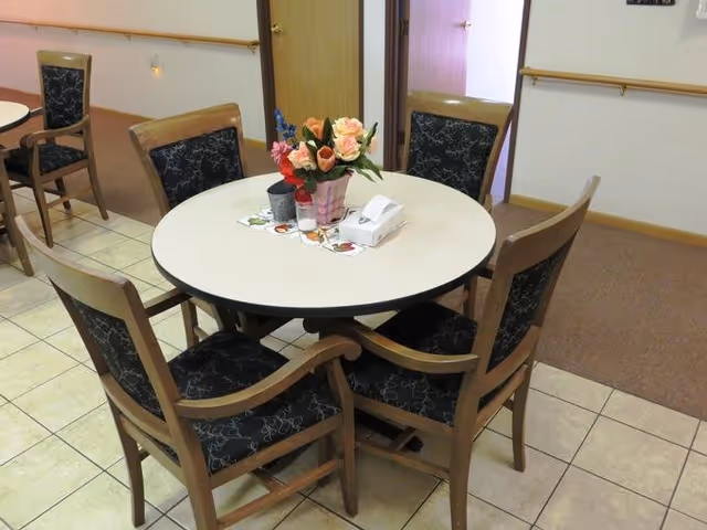 A round white table surrounded by five wooden chairs with dark patterned cushions in a room with tiled flooring and beige walls. On the table, there is a floral centerpiece with pink and red flowers, a tissue box, and some small items. Two open doors are visible in the background.