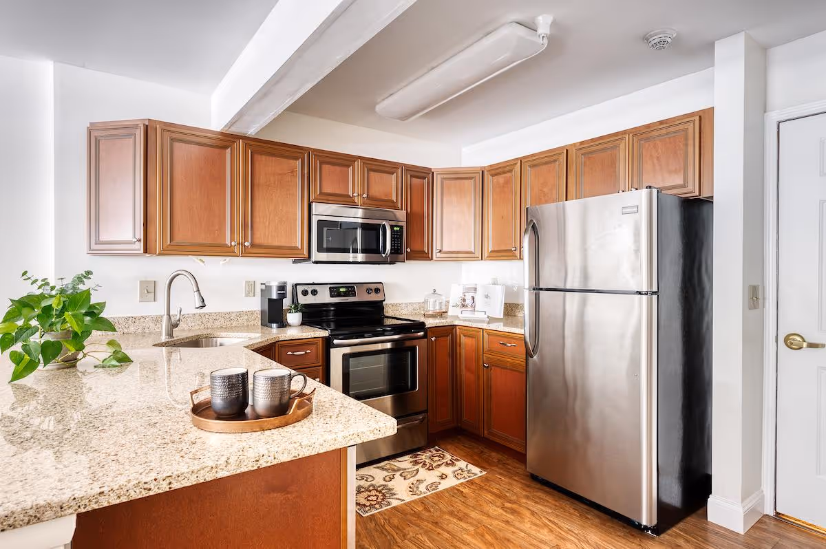 A modern kitchen with wooden cabinets, granite countertops, a stainless steel refrigerator, oven, and microwave. There is a small plant on the counter and two mugs on a tray. The floor is wooden, and the walls are white.