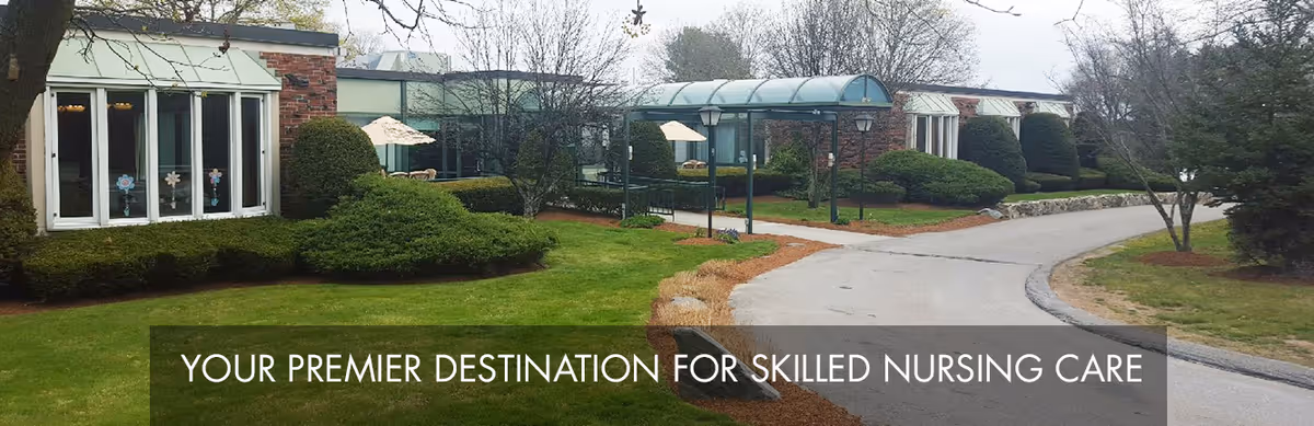 Exterior view of a single-story brick building with large windows, manicured bushes, trees, and a curved driveway. There is a covered walkway with a glass roof and outdoor umbrellas visible in the garden area.
