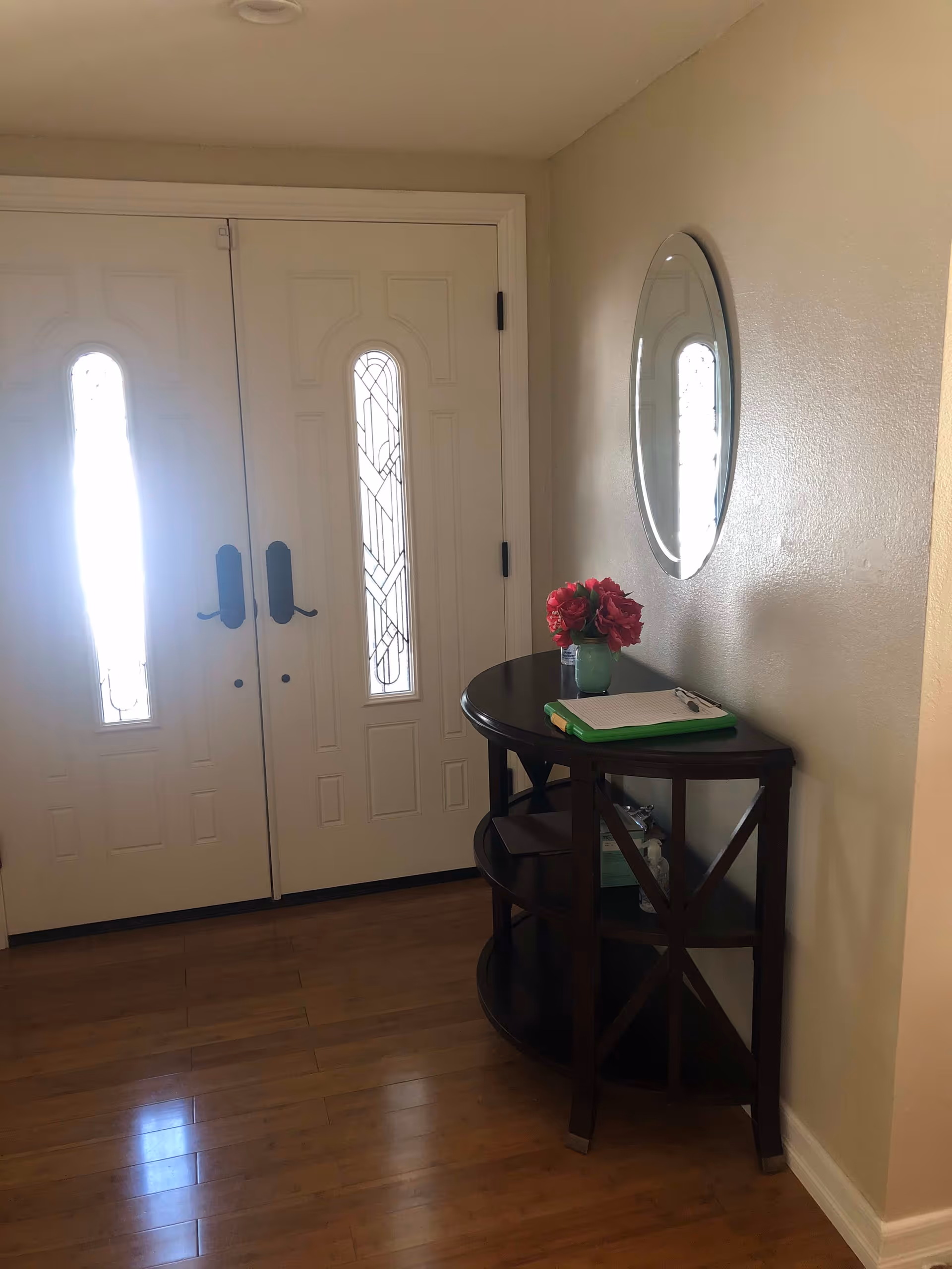 Entryway with double white doors featuring vertical decorative glass panels. A dark wooden half-moon console table is placed against the right wall, holding a green vase with red flowers and a clipboard with papers. Above the table is an oval mirror reflecting part of the door's glass panel. The floor is wooden and the walls are painted beige.