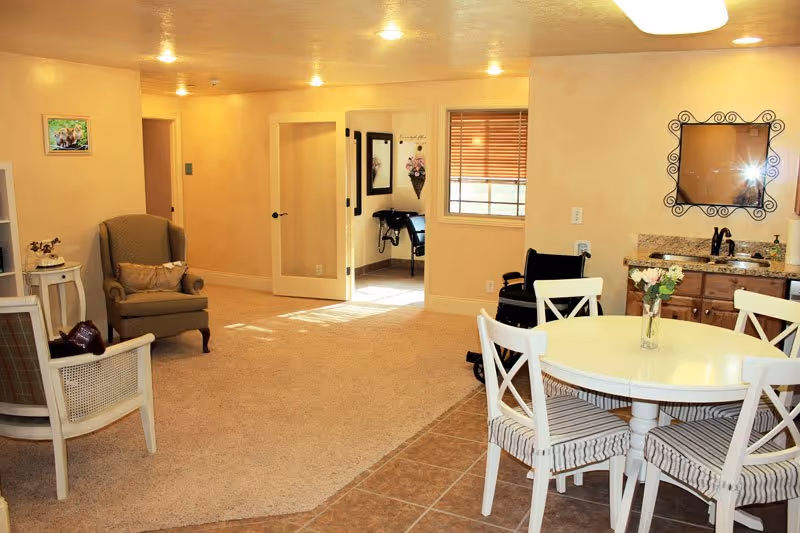 Bright assisted living common area with a round dining table and chairs, armchairs, a small sink area, and a wheelchair near an open doorway.