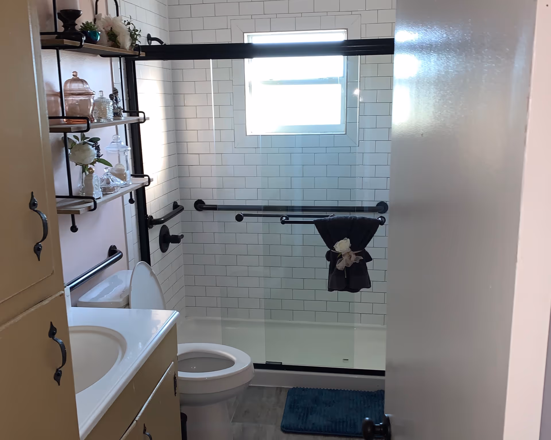 Small bathroom with a sink and toilet in front of a glass-enclosed subway-tiled shower fitted with grab bars and decorative shelving.