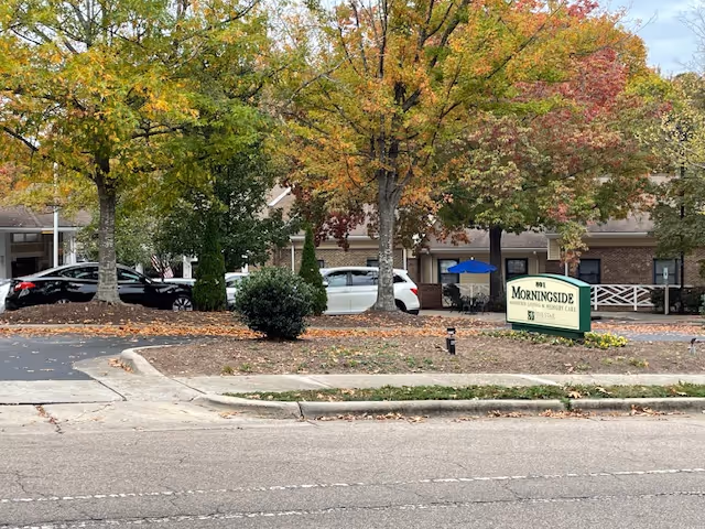 Front view of Morningside of Raleigh assisted living building with trees, parked cars, and a green sign on the lawn.