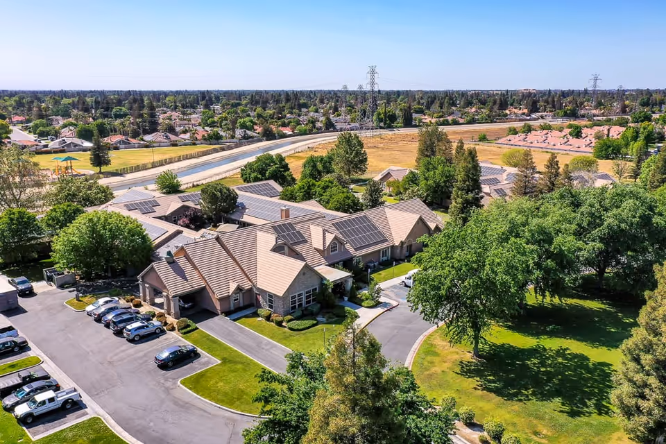 Aerial view of Riverstone Terrace Senior Living facility showing a large building with multiple roof sections, surrounded by green trees and lawns. There is a parking lot with several cars parked, and residential neighborhoods and open fields are visible in the background under a clear sky.