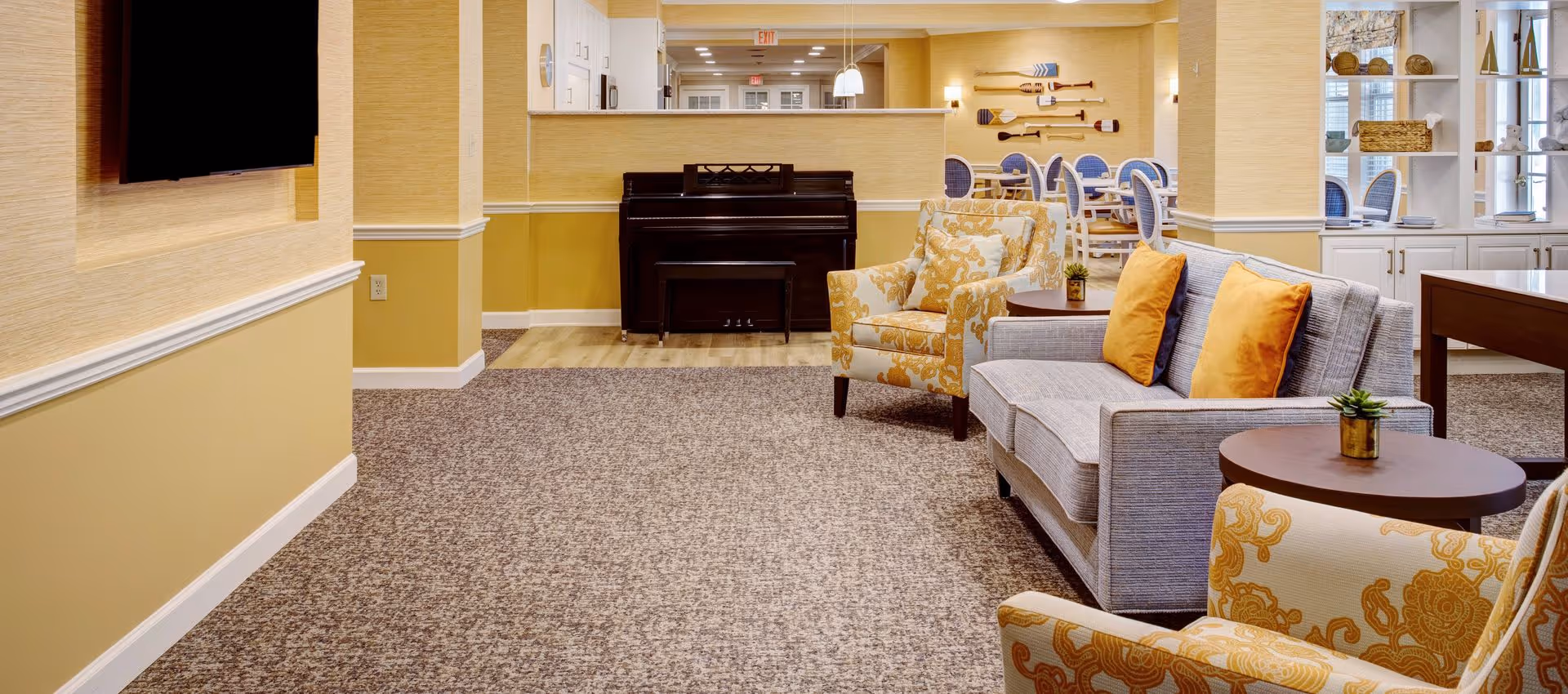 A cozy senior living facility common area with a gray sofa adorned with orange pillows, two patterned armchairs, a small round wooden table with a small plant, a piano against the wall, and a dining area with white chairs and blue cushions in the background. The walls are painted yellow with white trim, and there is a flat-screen TV mounted on the wall.