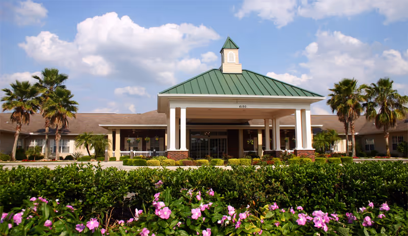 Front exterior view of Hawthorne Inn of Lakeland assisted living and memory care facility with a green roof, white pillars, palm trees, and flowering bushes in the foreground under a partly cloudy sky.