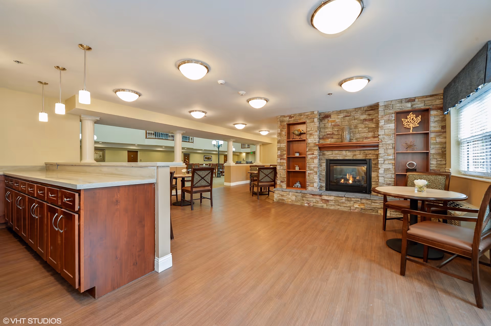 A spacious common area with wooden flooring featuring a stone fireplace with built-in shelves on either side. There are several round tables with chairs around them, and a countertop with wooden cabinets underneath. The room is well-lit with ceiling lights and pendant lights, and a window with blinds is visible on the right side.