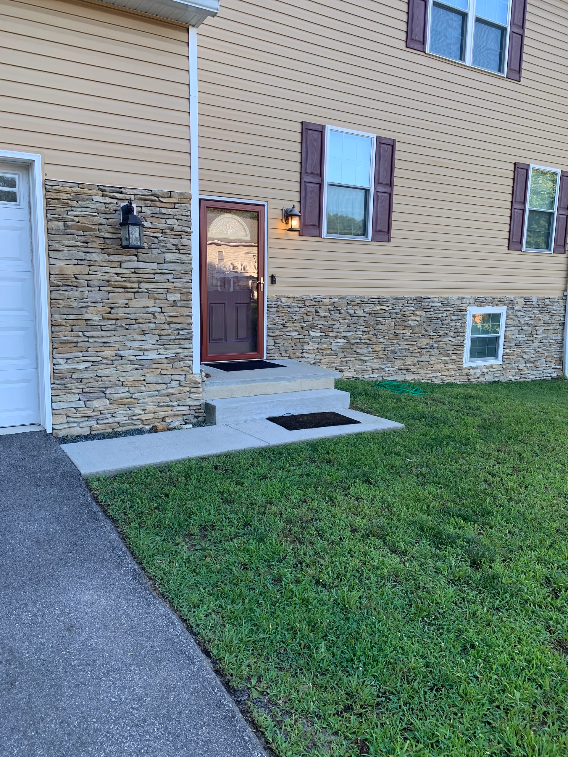 Front entrance of a beige siding house with stone veneer, a red door, concrete steps, exterior lights, and a grassy lawn.