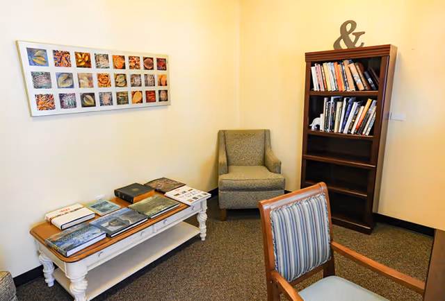A small reading or sitting area with a cushioned armchair, a wooden bookshelf filled with books, a wooden table with several books and magazines on it, and a striped cushioned chair in the foreground. The walls are light-colored and there is a framed artwork with multiple small images hanging on the wall.