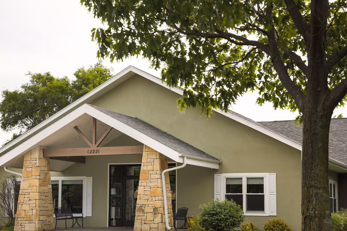 Front exterior of a single-story assisted living building with a covered stone-pillared entrance and a large tree in the foreground.