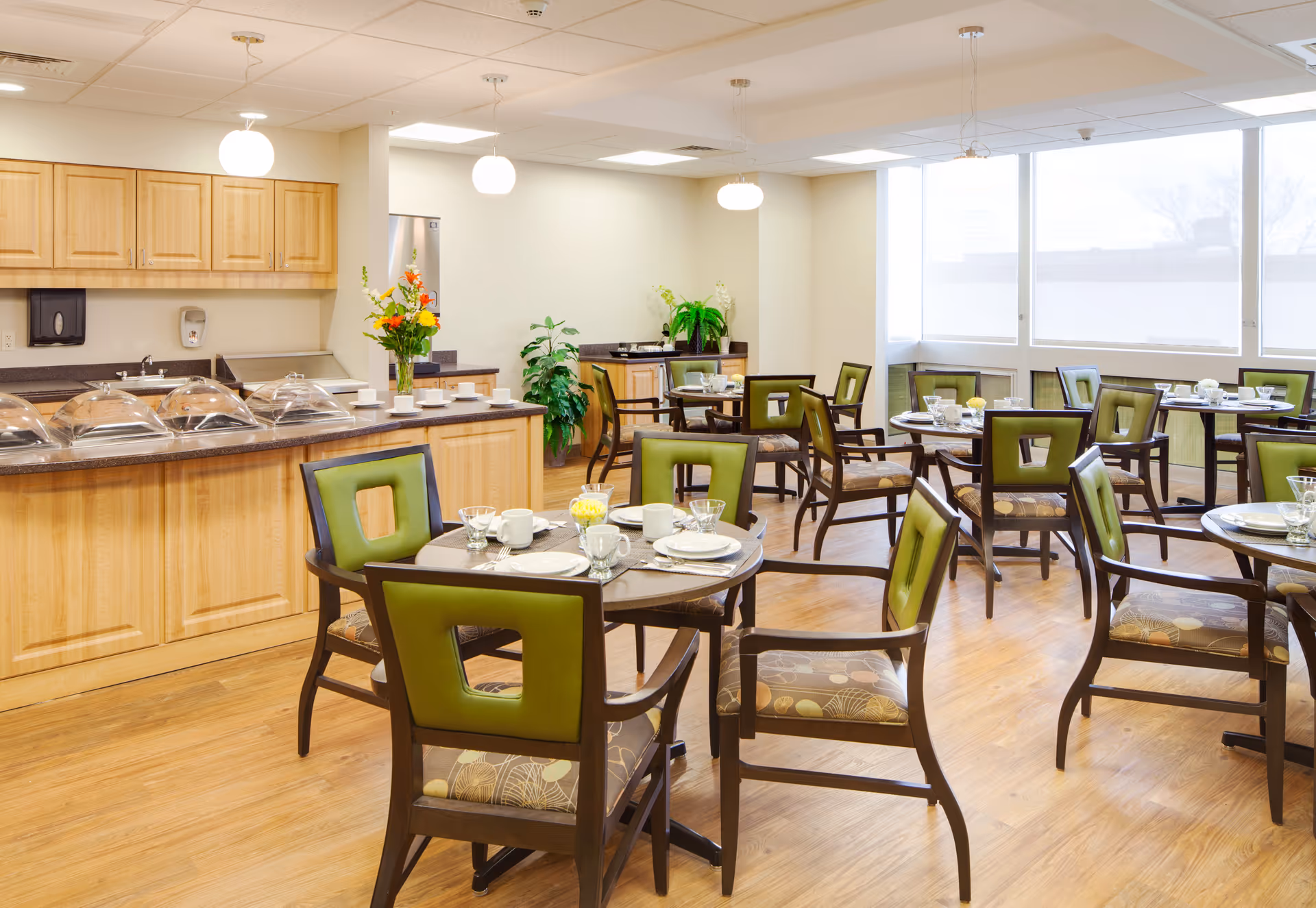 A bright dining room with several round tables set with white plates, cups, and glasses. Each table is surrounded by wooden chairs with green cushioned backs and patterned seats. The room features wooden flooring, large windows letting in natural light, and a serving counter with covered food trays and a vase of flowers. Potted plants are placed around the room, creating a welcoming atmosphere.