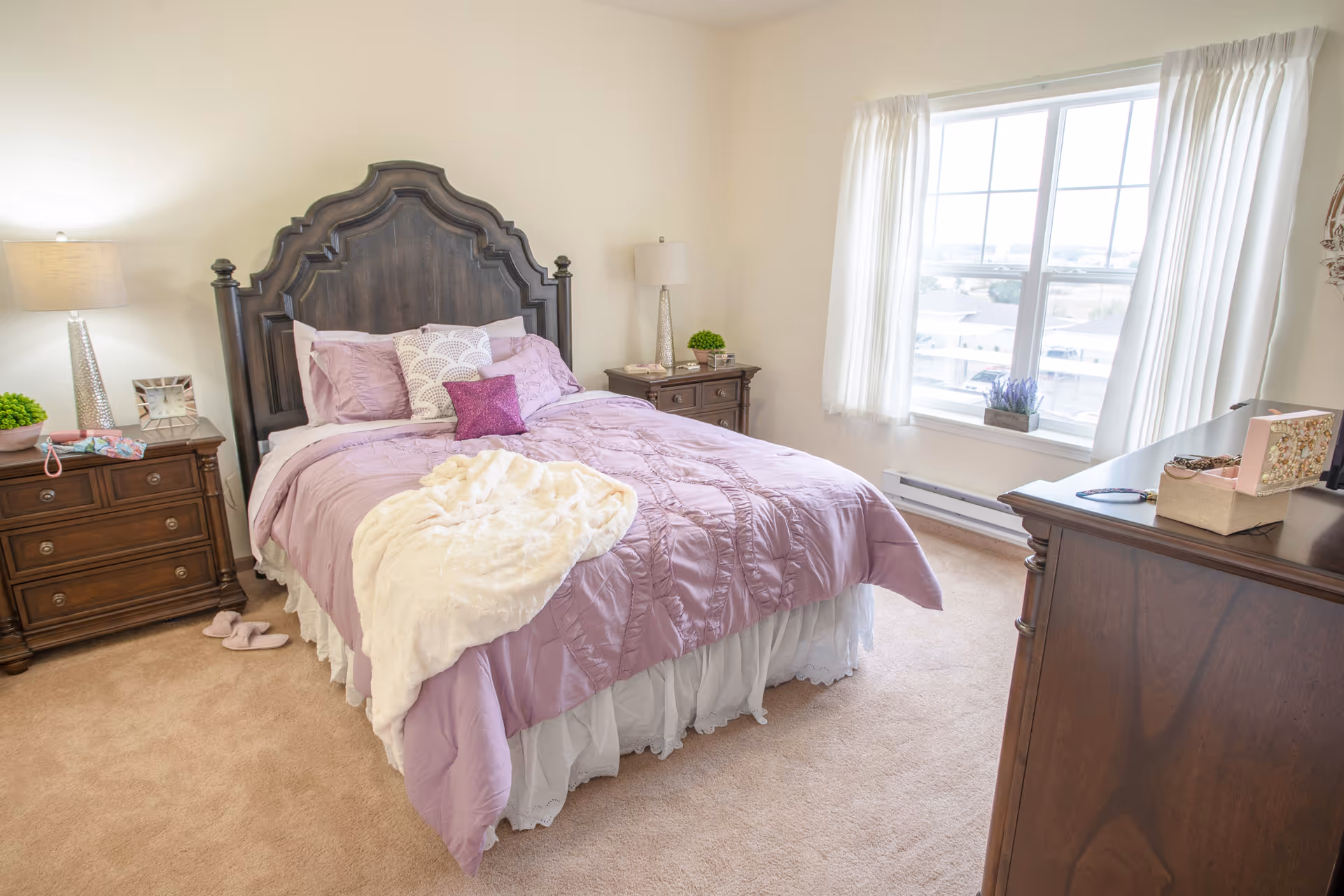 Sunlit bedroom featuring a large dark wood bed with lavender bedding, two nightstands, a dresser, and a window with white curtains.