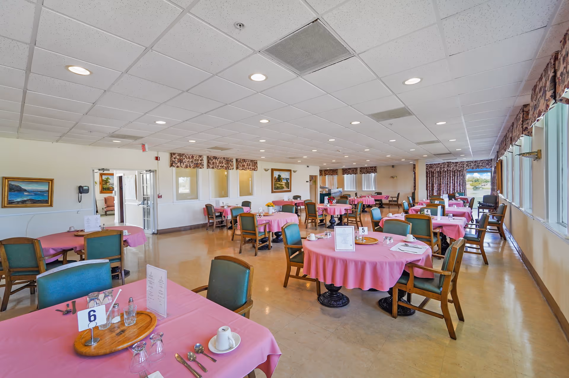 A spacious dining room with multiple round tables covered in pink tablecloths, each set with utensils, glasses, and menus. The room has green cushioned chairs, large windows with floral valances letting in natural light, and paintings on the walls. The floor is a light tan color and the ceiling has recessed lighting.