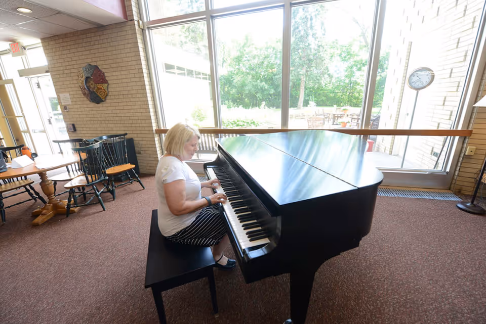 A woman with blonde hair wearing a white shirt and striped skirt is sitting on a bench playing a black grand piano in a room with large windows showing greenery outside. The room has carpeted floors, wooden chairs, and a round wooden table near a brick wall with a decorative wall hanging.