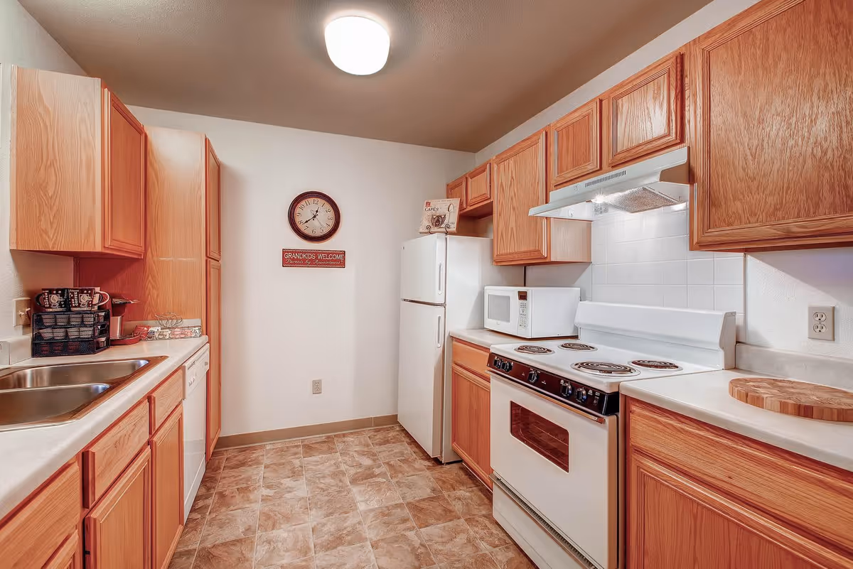 A kitchen with wooden cabinets, a white refrigerator, a white electric stove with an oven, a white microwave, a double sink, and a dishwasher. A clock and a sign that reads 'GRANDKIDS WELCOME' are mounted on the wall. The floor has a tile pattern, and the ceiling has a round light fixture.