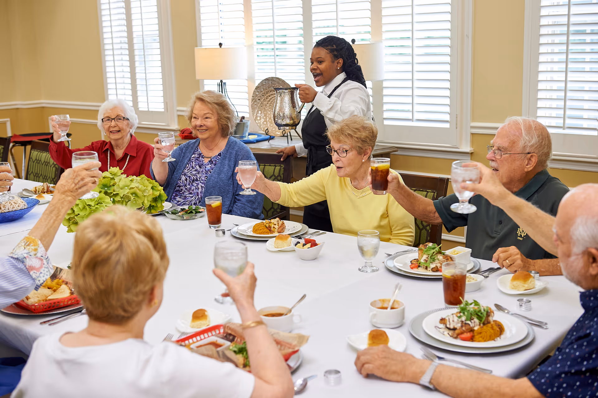 A group of elderly people sitting around a dining table raising their glasses in a toast while a server stands nearby holding a pitcher. The table is set with plates of food, glasses of water and iced tea, and rolls. The room has large windows with white shutters and a lamp in the background.