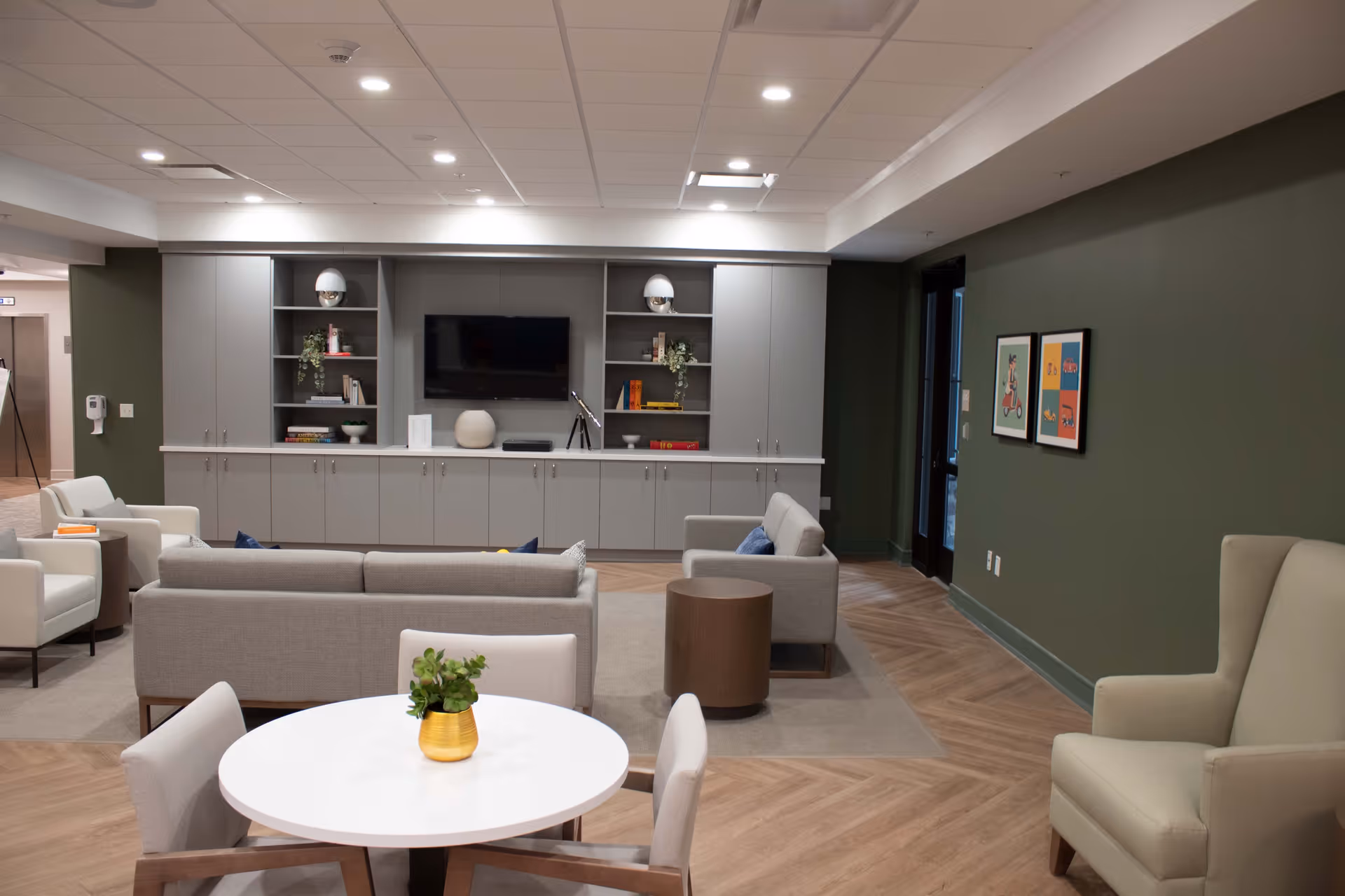A modern and cozy living room area in a senior living facility with a round white table and four chairs in the foreground, a gray sofa and armchairs arranged around a small round wooden table, built-in gray cabinets and shelves with decorative items, a flat-screen TV mounted on the wall, green painted walls, wooden flooring, and framed artwork on the wall.