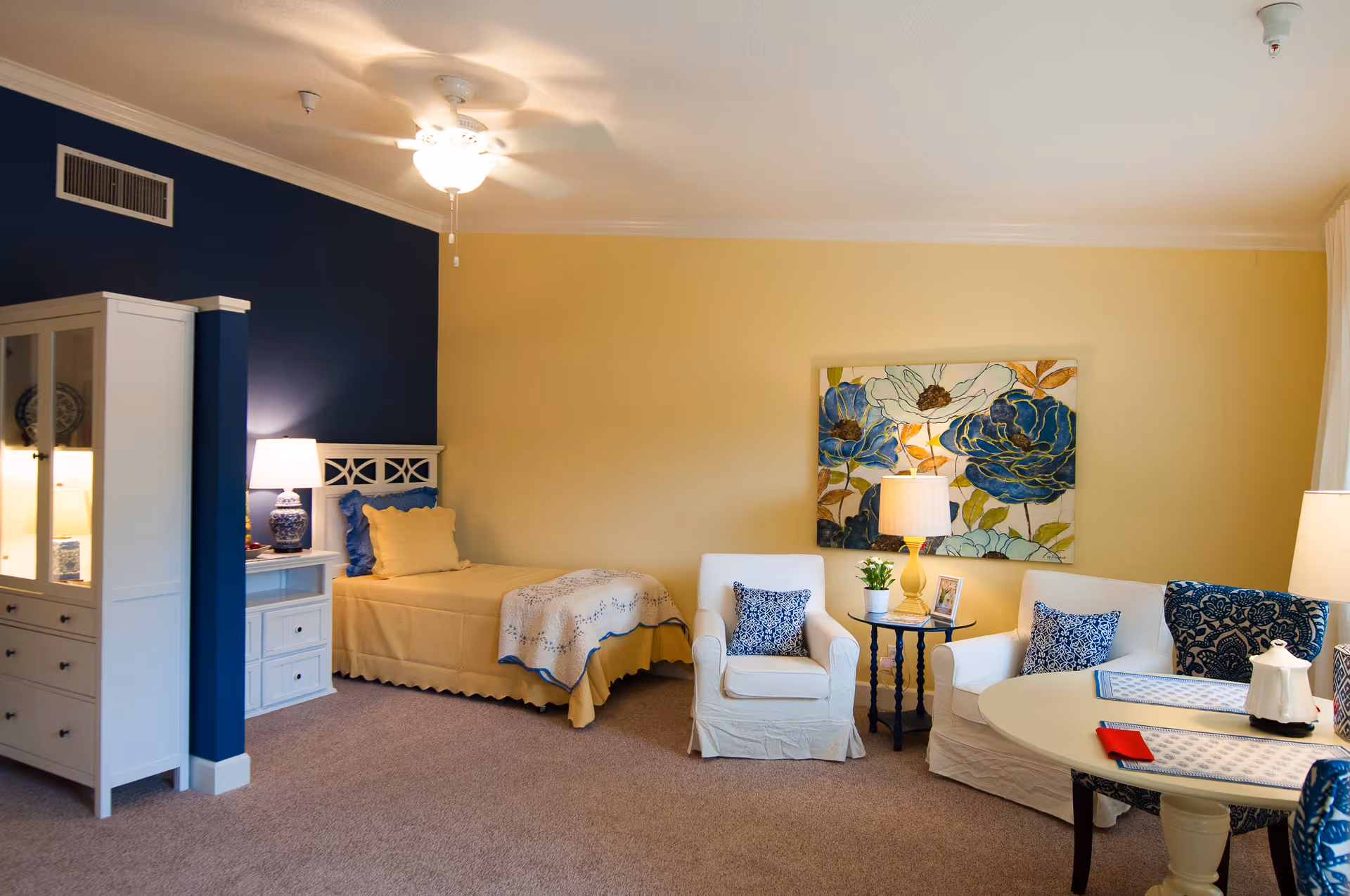A cozy senior living room with a single bed against a blue accent wall, a white nightstand with a lamp, a white cabinet, two white armchairs with blue patterned pillows, a round table with placemats, and a floral painting on a yellow wall.