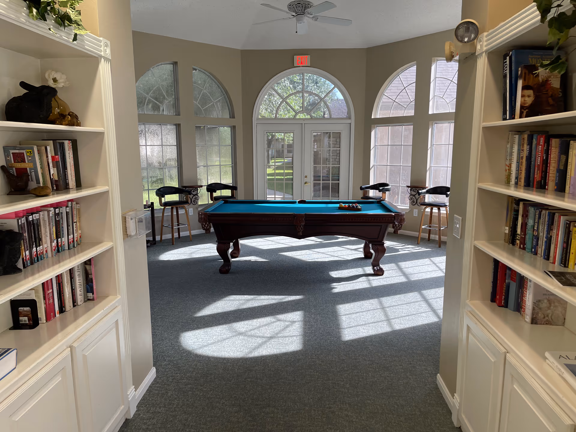 A bright room with large arched windows and a glass door letting in sunlight. In the center is a pool table with a set of billiard balls arranged on it. Four bar stools with black seats are positioned around the pool table near the windows. On either side of the entrance to the room are built-in white bookshelves filled with books and decorative items.