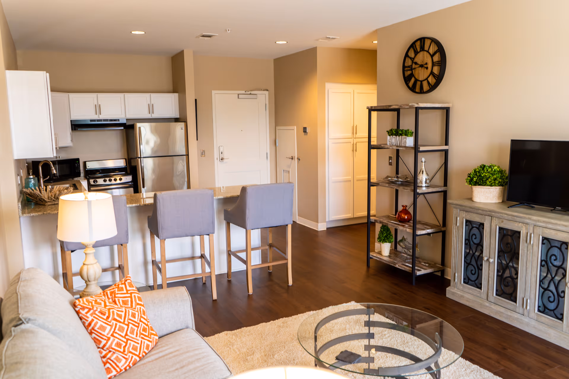 A modern open-concept living area featuring a kitchen with white cabinets, stainless steel appliances, and a granite countertop with three gray bar stools. The living room includes a beige sofa with an orange patterned pillow, a glass coffee table on a light rug, a wooden TV stand with a flat-screen TV, a black metal shelving unit with decorative items, and a large wall clock. The space has warm beige walls and dark wood flooring.