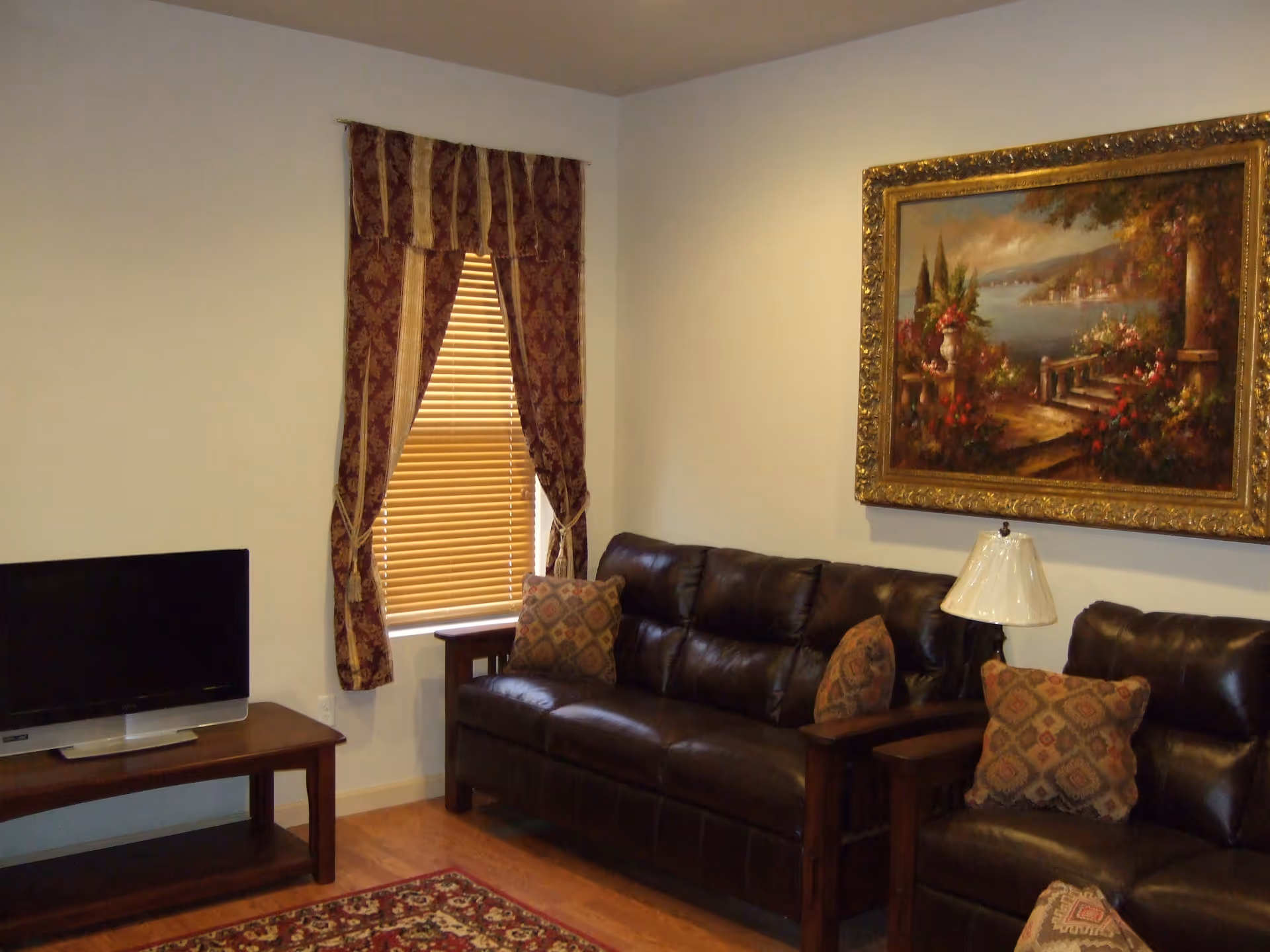 A cozy living room with two dark brown leather sofas adorned with patterned cushions. A wooden side table with a lamp is placed between the sofas. A flat-screen TV sits on a wooden TV stand to the left. The window has wooden blinds and is framed by decorative maroon and gold curtains. A large framed painting depicting a scenic landscape with a garden and water view hangs on the wall above the sofas.