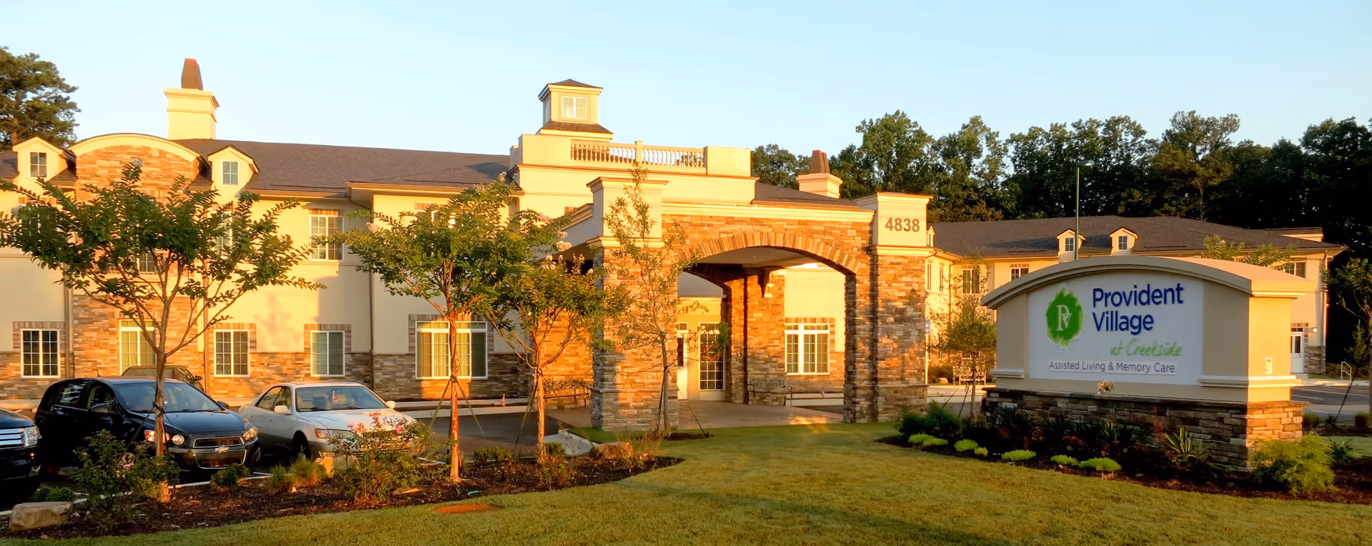 Exterior view of a senior living facility building with a stone and beige facade, several windows, and an arched entrance. There are trees, landscaped plants, and parked cars in front. A large sign in the foreground reads 'Provident Village at Creekside Assisted Living & Memory Care'.