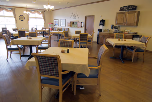Dining room with several tables and chairs arranged on wood flooring, a beverage station and cabinets along the back wall, and large windows letting in daylight.