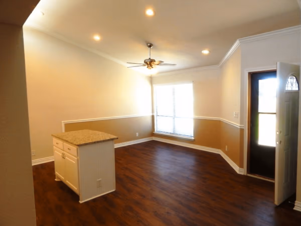 Empty open living area with hardwood floors, a small kitchen island, ceiling fan, large window with blinds, and an open front door.