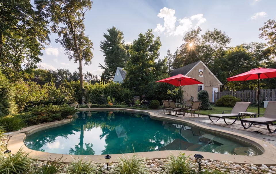Outdoor swimming pool surrounded by greenery and trees with a small building in the background. There are patio chairs and tables with red umbrellas around the pool area under a partly cloudy sky with the sun shining through the trees.
