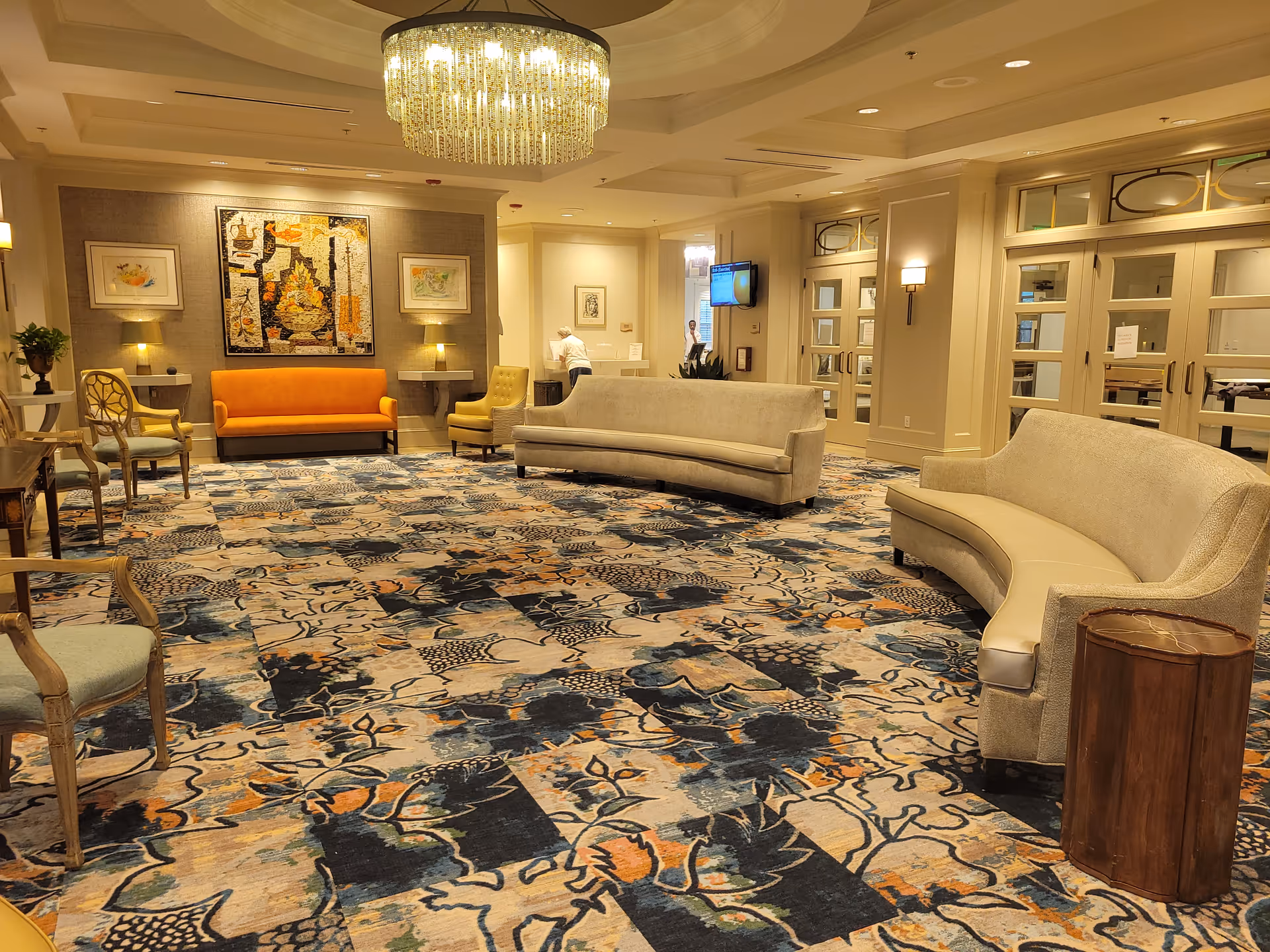 Bright, elegant senior living lobby with patterned carpet, curved sofas and chairs, a chandelier, and wall artwork.