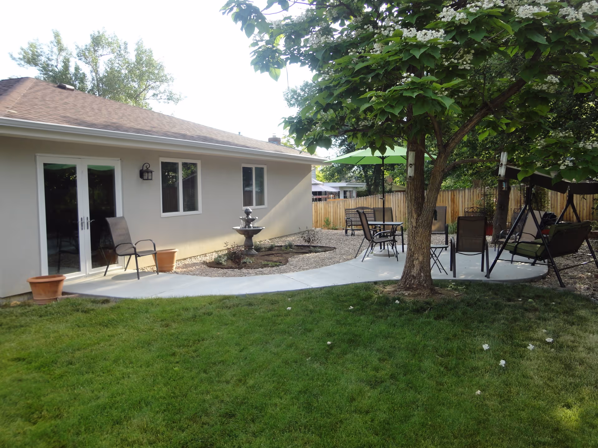 Outdoor garden area at Peak Gardens Assisted Living featuring a green lawn, a concrete pathway, a tree with green leaves, patio furniture including chairs, tables with umbrellas, a swing, and a small water fountain near the building wall.