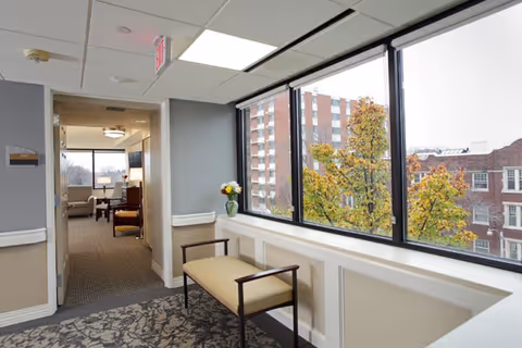 Interior hallway area in a senior living facility with large windows showing an outside view of buildings and trees. A bench with armrests is placed against the wall under the windows, and a vase with flowers is on the windowsill. The hallway leads to a common area with chairs and a table visible in the background.