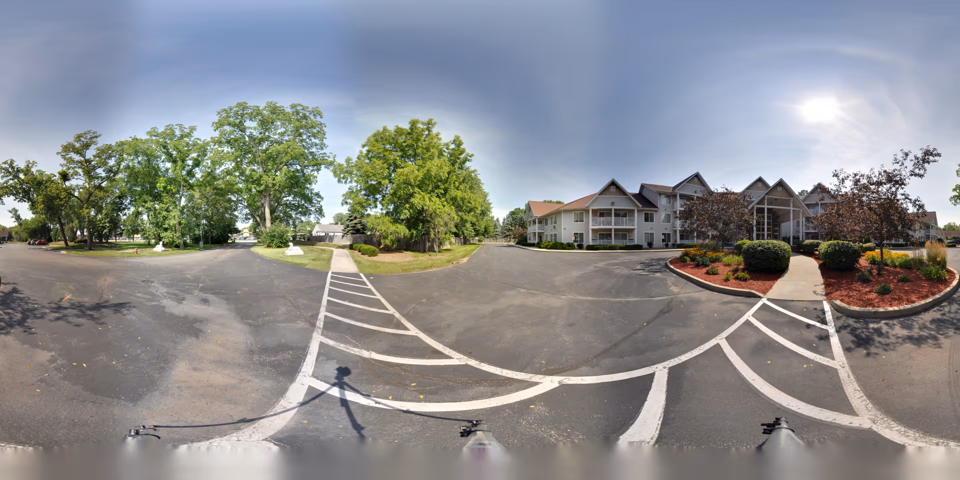 Wide panoramic view of the exterior of Charter Senior Living of Kenosha, showing a large parking area, landscaped garden beds with bushes and small trees, and a multi-story building with balconies under a partly cloudy sky.