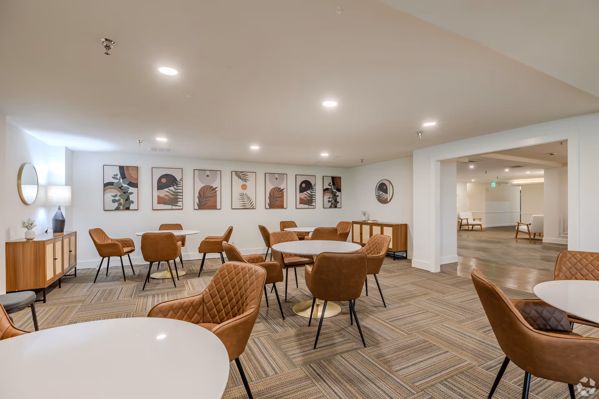Interior view of a communal dining or meeting area in a senior living facility, featuring round white tables surrounded by brown cushioned chairs with quilted backs. The room has a patterned carpet, white walls adorned with modern abstract artwork, and wooden sideboards with decorative items. The space is well-lit with ceiling lights, and an adjacent room with additional seating is visible through a large open doorway.