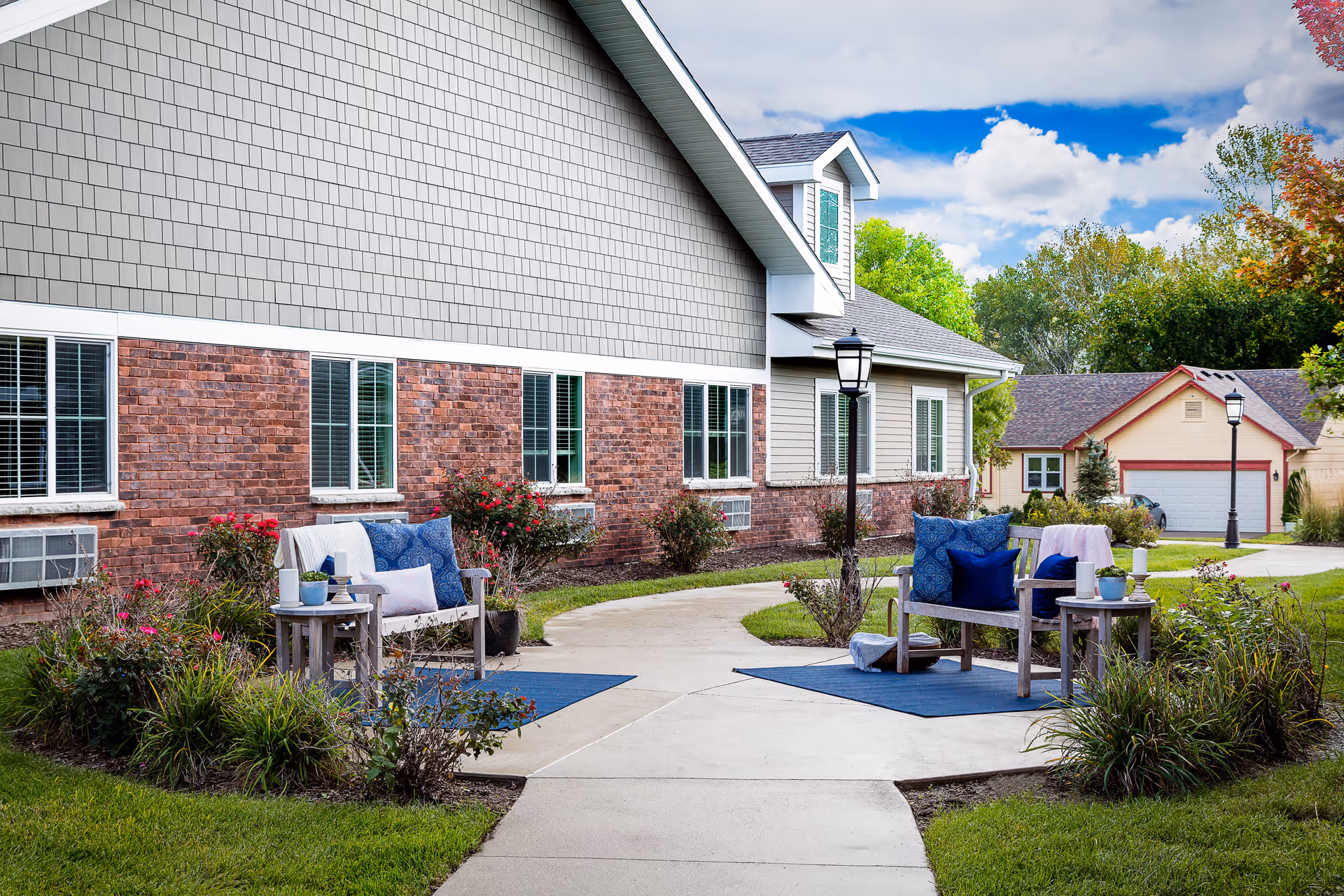 Outdoor seating area with two wooden benches adorned with blue and white cushions, small side tables with potted plants, and a concrete pathway leading past a brick and siding building. There are street lamps, green grass, bushes, and trees under a partly cloudy sky.