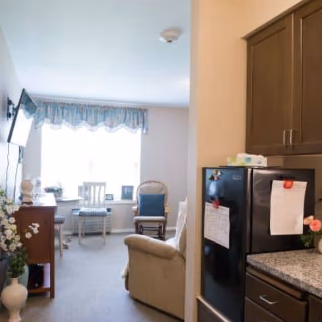 Interior view of a senior living facility room showing a small living area with a beige armchair, a wooden rocking chair with a blue cushion, two white chairs near a window with blue valance curtains, a wall-mounted TV, and a kitchen area with dark wood cabinets and a black refrigerator with papers attached.
