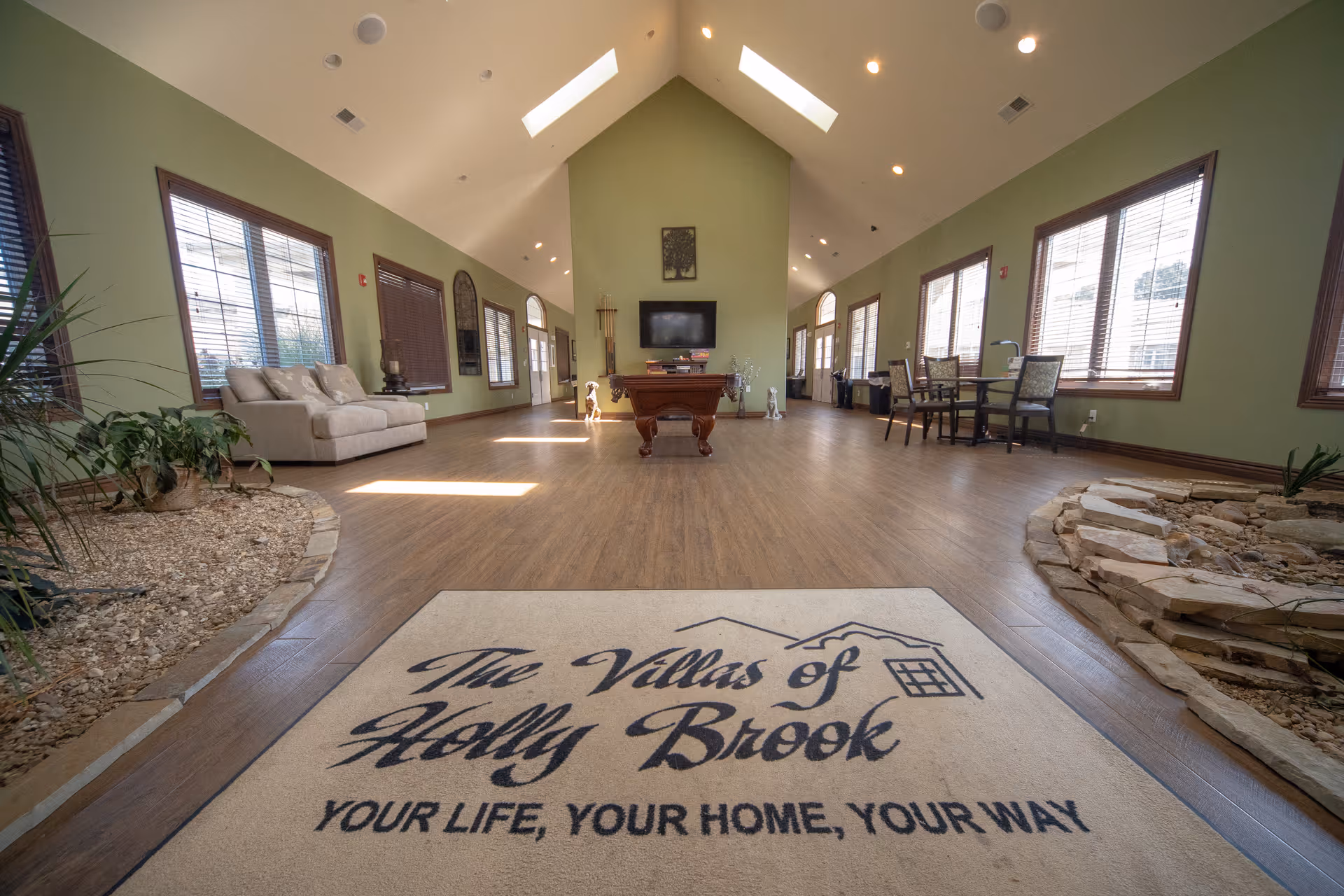 Spacious interior common area with high vaulted ceilings and multiple windows letting in natural light. The room features a pool table in the center, a beige couch on the left, and a small dining table with chairs on the right. There are indoor garden beds with rocks and plants on both sides near the entrance. A large mat on the floor near the entrance reads 'The Villas of Holly Brook YOUR LIFE, YOUR HOME, YOUR WAY.'