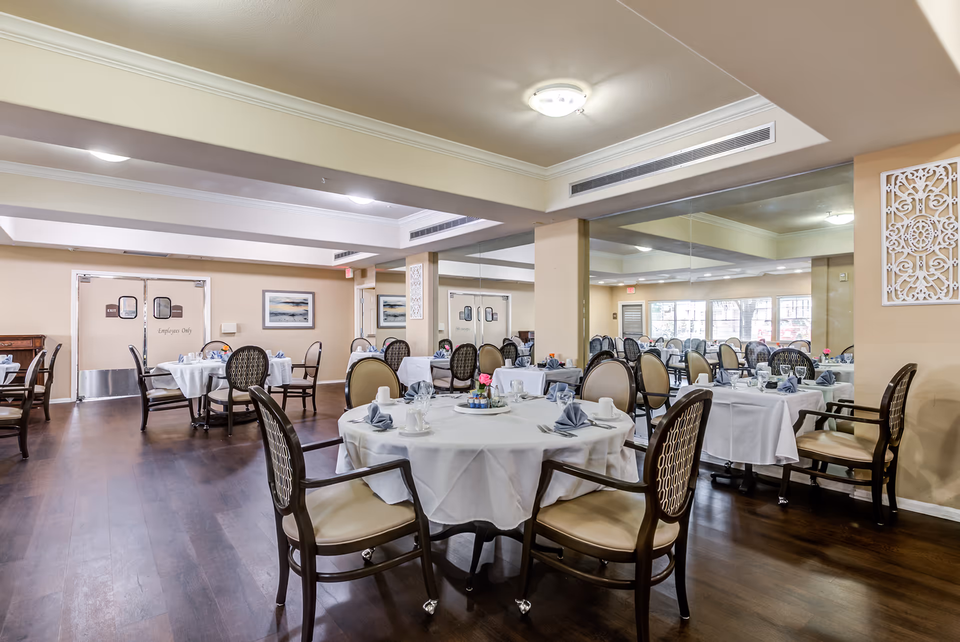 A spacious dining room with multiple round tables covered with white tablecloths, each set with folded napkins, cups, and small flower centerpieces. The room has wooden flooring, beige walls, and large mirrors on one side reflecting the tables and chairs. There are double doors labeled 'Employees Only' at the back of the room.