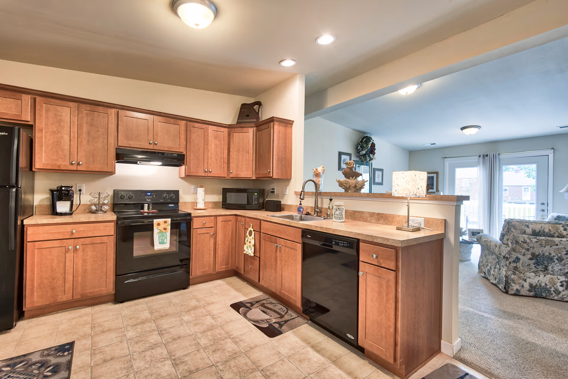 A kitchen with wooden cabinets, black appliances including a refrigerator, stove, microwave, and dishwasher. The kitchen has a tiled floor and a countertop with a sink and a lamp. Beyond the kitchen is a living room area with floral upholstered furniture and a glass door leading outside.